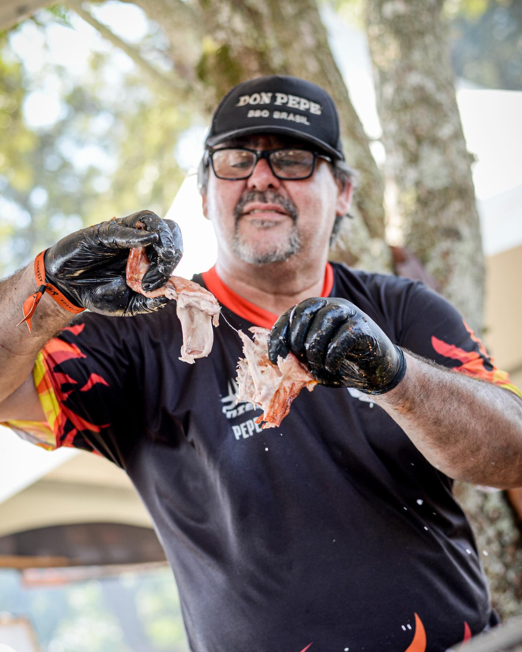 Don Pepe de luvas pretas e uniforme com chamas, desfiando carne na brasa durante um festival